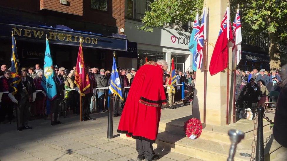 Man in Mayor's red robe and regalia bows having laid a wreath at the memorial. Several flags are visible