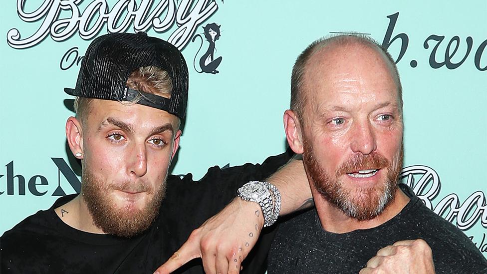 Jake Paul and his father Greg Paul looking away from the camera. The background is light blue with branding, Jake is wearing a cap backwards and a silver watch on his left hand, which is resting on Greg's right shoulder. They are both wearing dark tops.