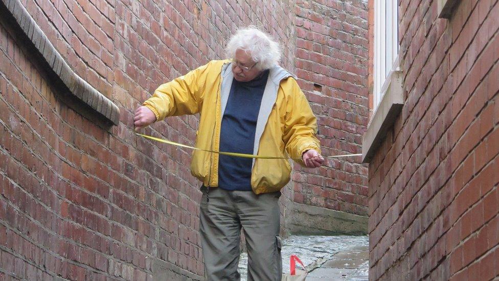 A man in a yellow coat measures the width of the alleyway