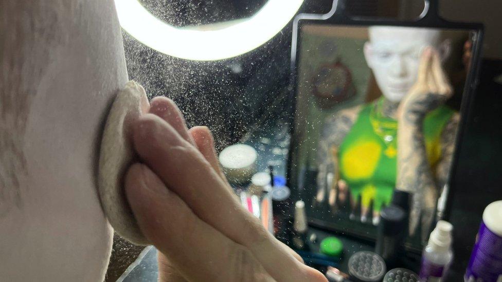 Danya applying white powder on his face in front of a mirror