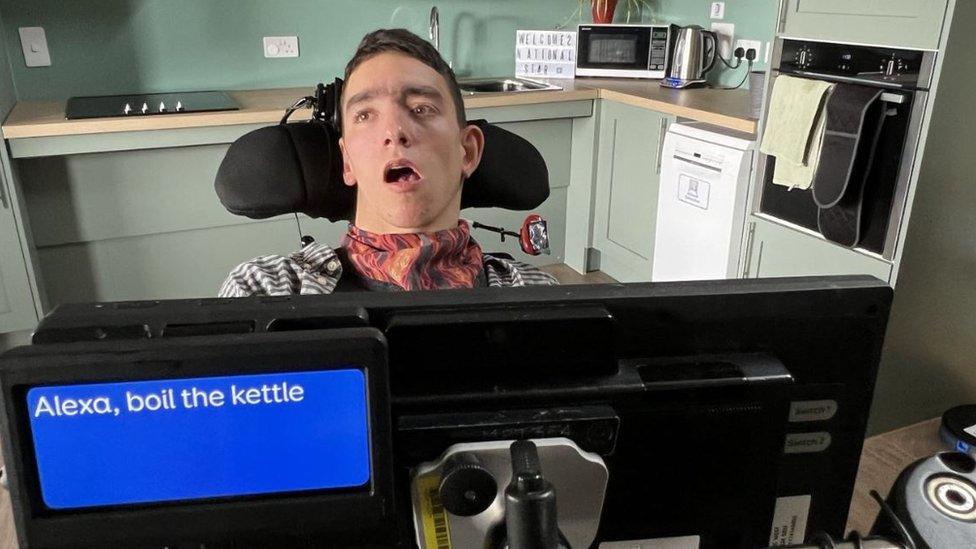 Jaspar sitting in the kitchen in his wheelchair, his electronic communicator is in front of him. It says 'Alexa boil the kettle' on the digital screen.