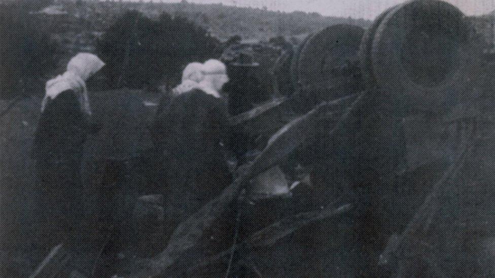 A black and white photo of women surrounding upturned remnants of a bus