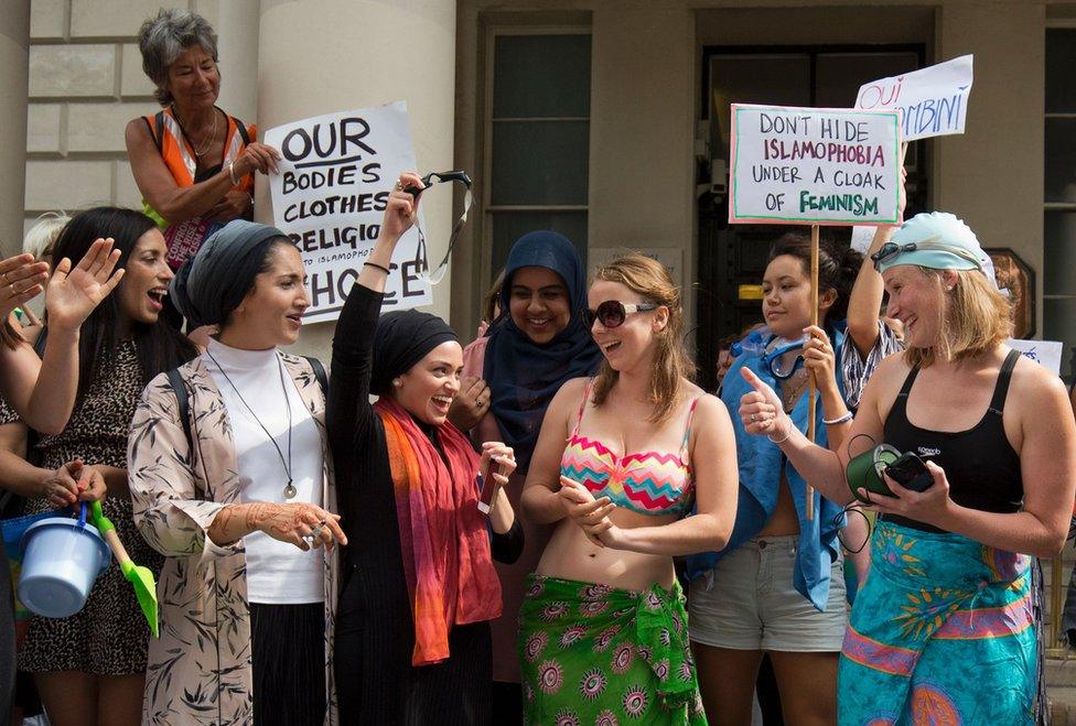 women in loads of different types of clothes (one in a bikini, some in head coverings) laugh and clap together