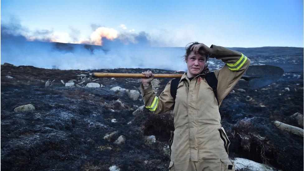 Hayley Agnew standing in a similar pose, amid the heat of the charred remains holding a shovel on her back, with smoke rising off of the Mourne Mountains behind her.