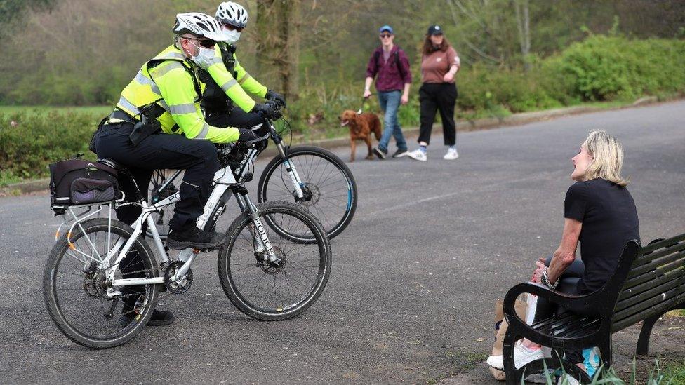 Police on bicycles talk to lady on bench