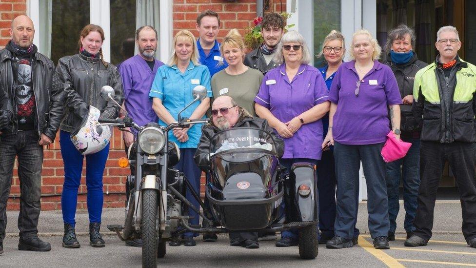 Joe Canning wearing sunglasses and sitting in a black motorcycle sidecar surrounded by care home staff and people wearing motorcycle gear