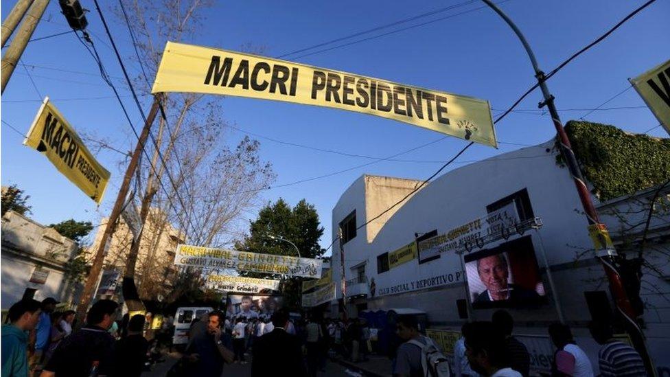 People walk past a TV screen where presidential candidate Mauricio Macri is seen outside a campaign rally in Lanus, on the outskirts of Buenos Aires on 21 October, 2015.