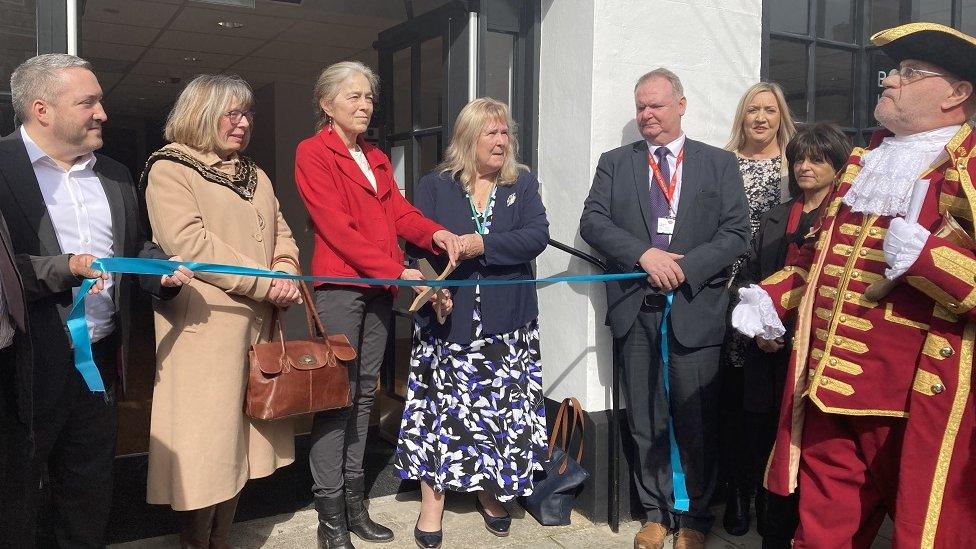 Dignitaries stand behind a blue ribbon in front of the hub.