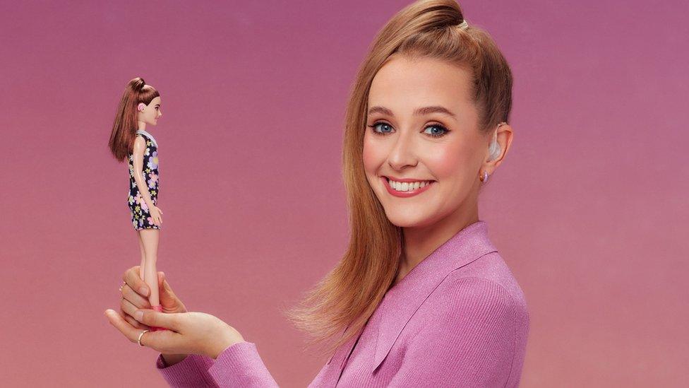 rose ayling-ellis holding a barbie doll with a hearing aid and smiling at the camera