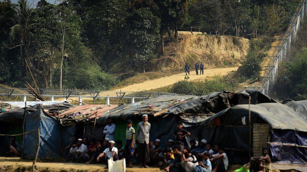 Myanmar security personnel keeps watch along the Myanmar-Bangladesh border as Rohingya refugee sit outside their makeshifts shelters near Tombru