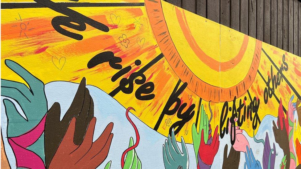 Mural outside the Harbour Project - hands reaching up towards a sun with the words "We rise by lifting others"