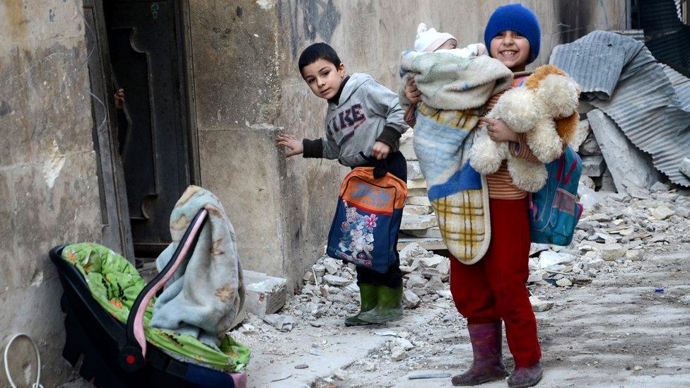 Syrian girl, holding her baby brother, stands next to the rubble