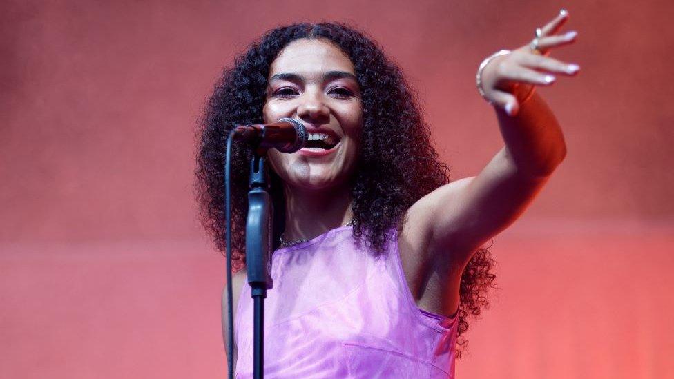 Olivia Dean performing on stage. The 24-year-old singer wears her dark curly hair loose and wears a purple short sleeved top and matching purple eye shadow. She smiles as she singes into a microphone on a stand in front of her, her left arm raised in front of her towards her audience. She's pictured on stage where there is a smoke machine lit by red lights