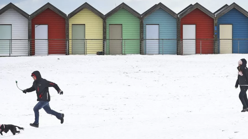 Colourful beach huts and people running with a dog on a snow covered beach in Northumberland