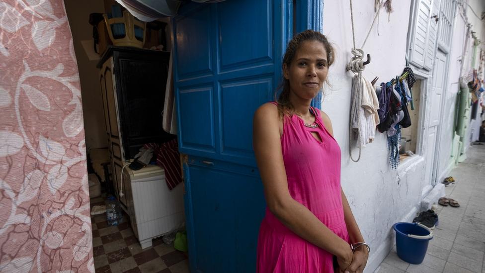 A woman dressed in pink stands leaned against a blue door