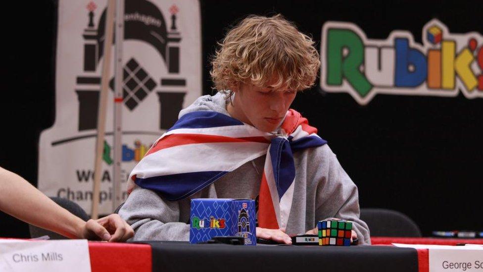 George Scholey with medium-length blond hair wearing a union flag cloak