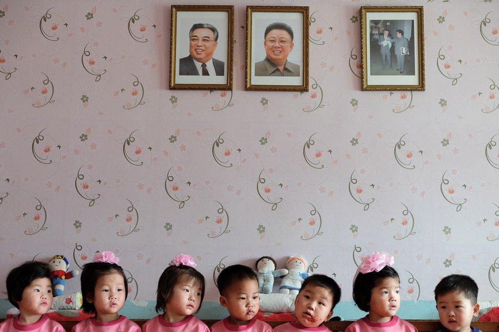 Young children in an orphanage sitting beneath portraits of Kim Il-sung and Kim Jong-il