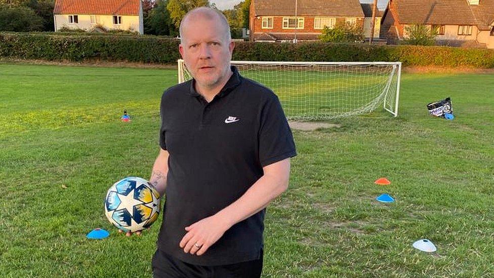 Simon Compton holding a football wearing a black T-shirt and black shorts