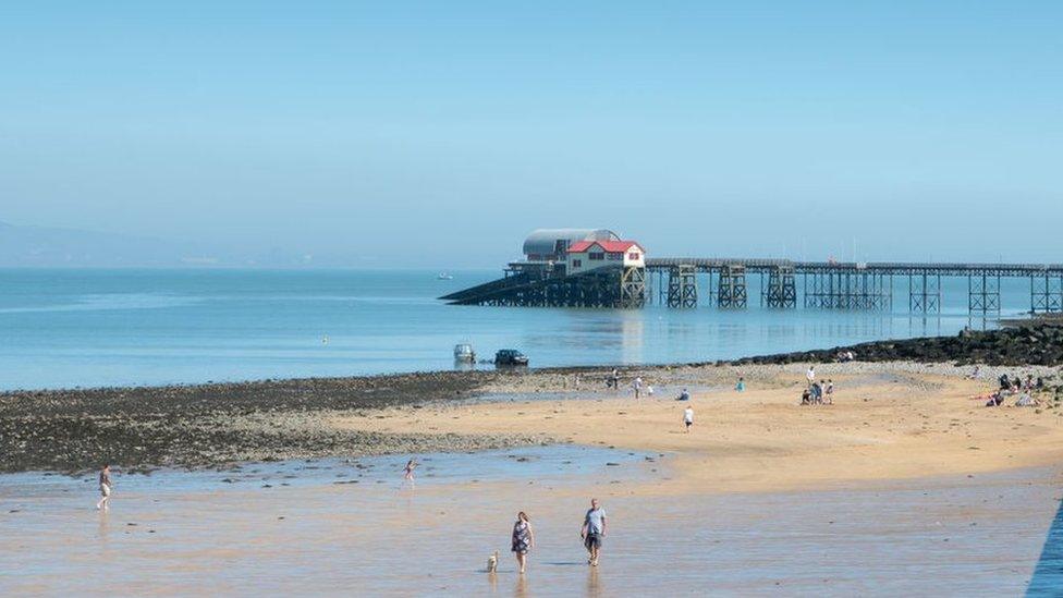 Mumbles, with pier in background and two people walking on beach, on a clear, bright day