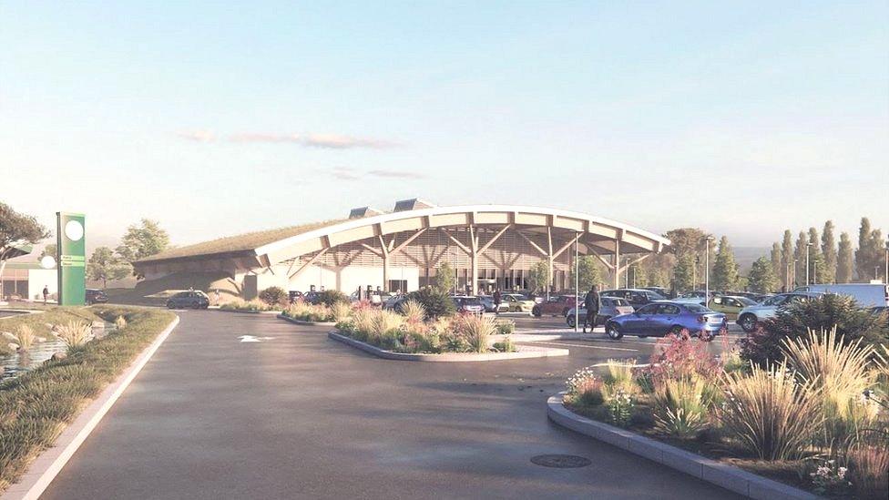 A proposed digital rendering of what the service station will look like. It has a rounded roof and lots of pillars holding it up, a large car park and surrounded by greenery