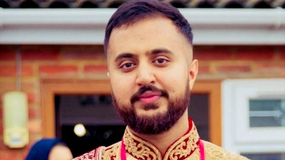 Mohammed standing in front of the back of a house. Faint bricks in the backgrounds. He is wearing his wedding shiwani which is white with red and gold embroidery.