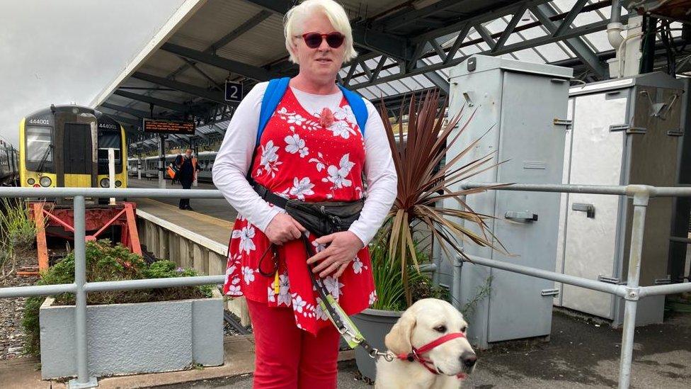 blind woman wearing red jeans standing with golden labrador guide dog at train station