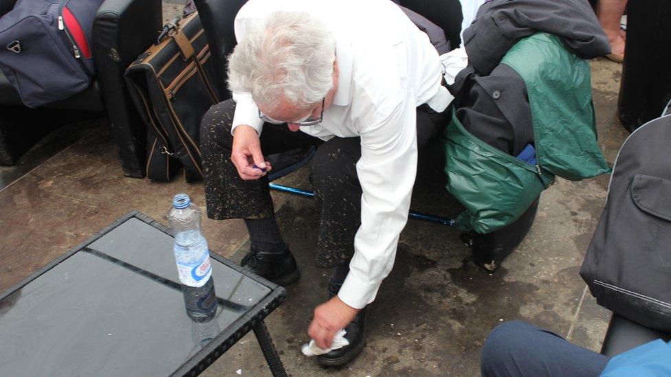 Aelod o gor yn sychu'r mwd o'i esgidiau // A choir member wiping the mud from his shoes