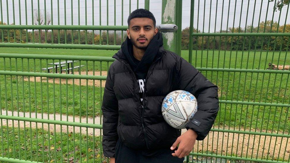 Harun Hamid standing in front of a green metal fence with a grass field behind him. He is British Pakistani and is looking at the camera. He has short black hair and a short black beard and moustache. He is wearing a black puffer jacket and has a black hoodie underneath it. He is holding a white football under his left arm.