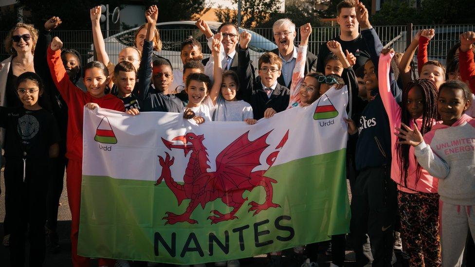 school children holding a Welsh flag with the Urdd symbol and name Nantes on it. Children are in front of some adults, including First Minister Mark Drakeford