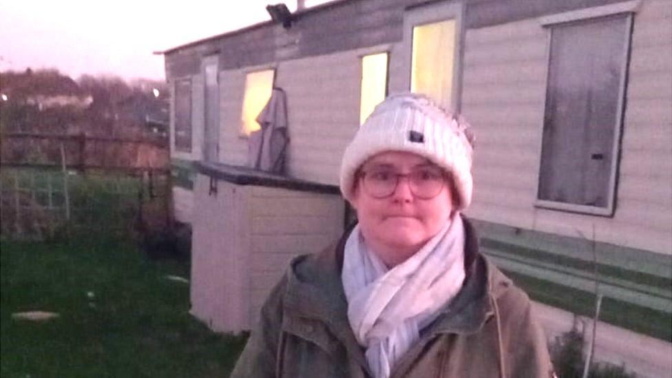 Jaimee Bremer wearing a hat, scarf and coat, stands outside her caravan in the early morning