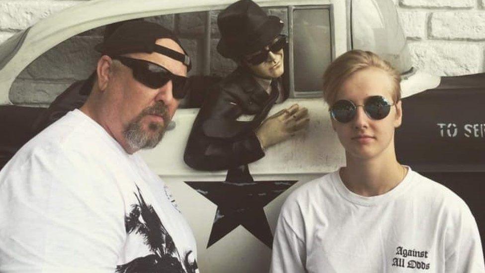 Gaby Hutchinson (right) poses in front of a Blues Brothers mural. Gaby, a white woman in her 20s, has short blonde hair - she wears round sunglasses and a white T-shirt and looks at the camera with a cool expression. She is pictured with her father who stands on her left - he has a grey beard and also wears sunglasses and a white T-shirt as well as a black baseball cap twisted backwards.