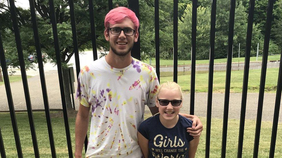 A young adult with bright pink hair smiles and hugs a little girl who is grinning broadly