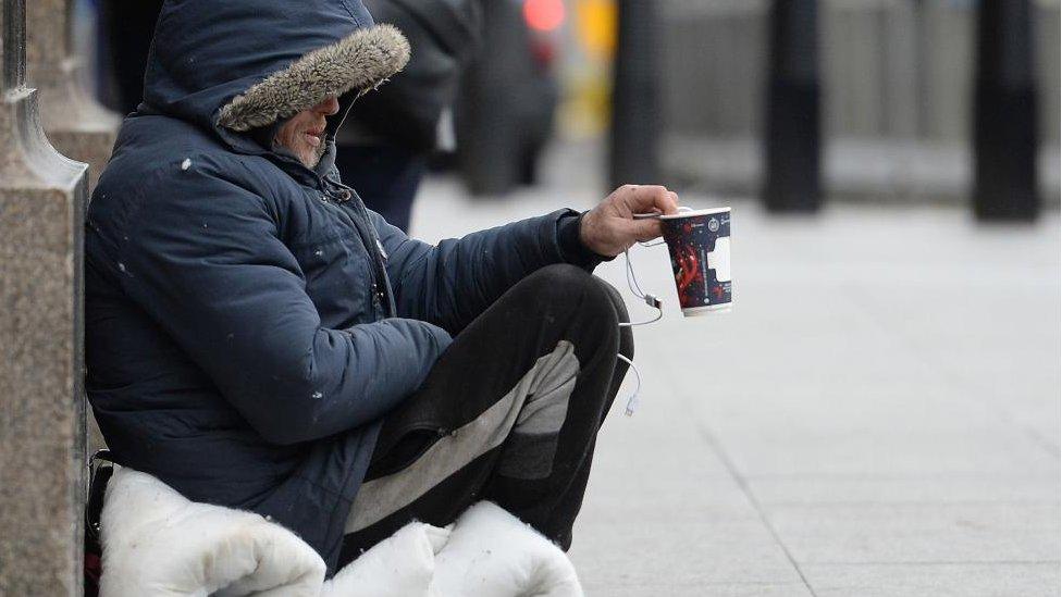 Man with face mostly hidden by large padded coat with hood holds out a paper cup to passers by