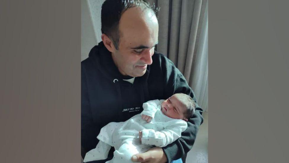 Raj Gill holding his newborn baby. He is wearing a black hoodie and smiling at his baby boy. The baby is wearing white.