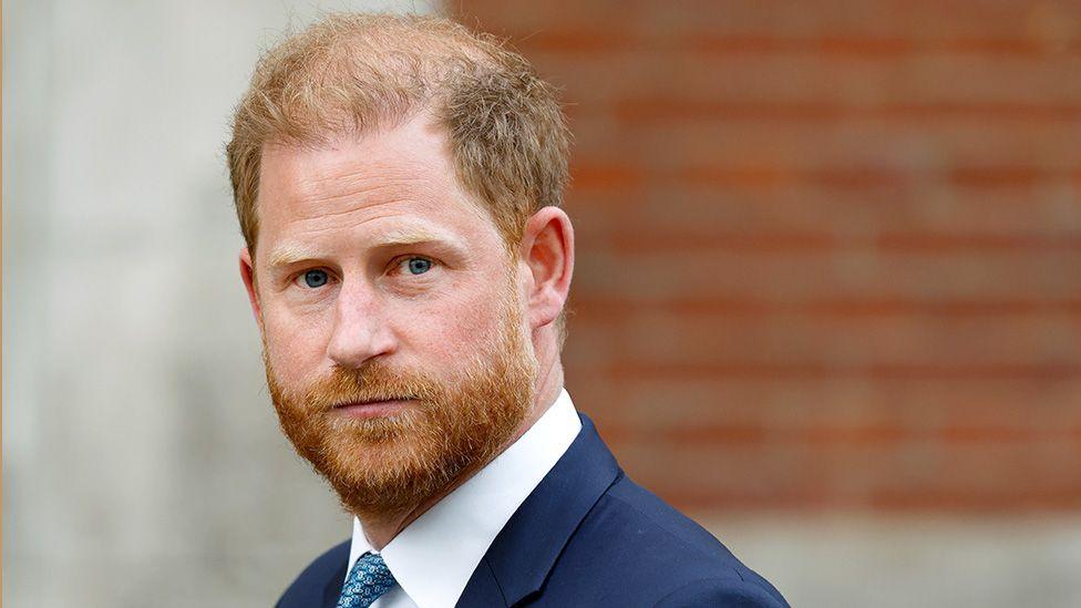 Prince Harry is wearing a dark suit jacket, white dress shirt, and blue patterned tie. The background consists of a brick wall on the right side and a light-colored concrete structure on the left side.