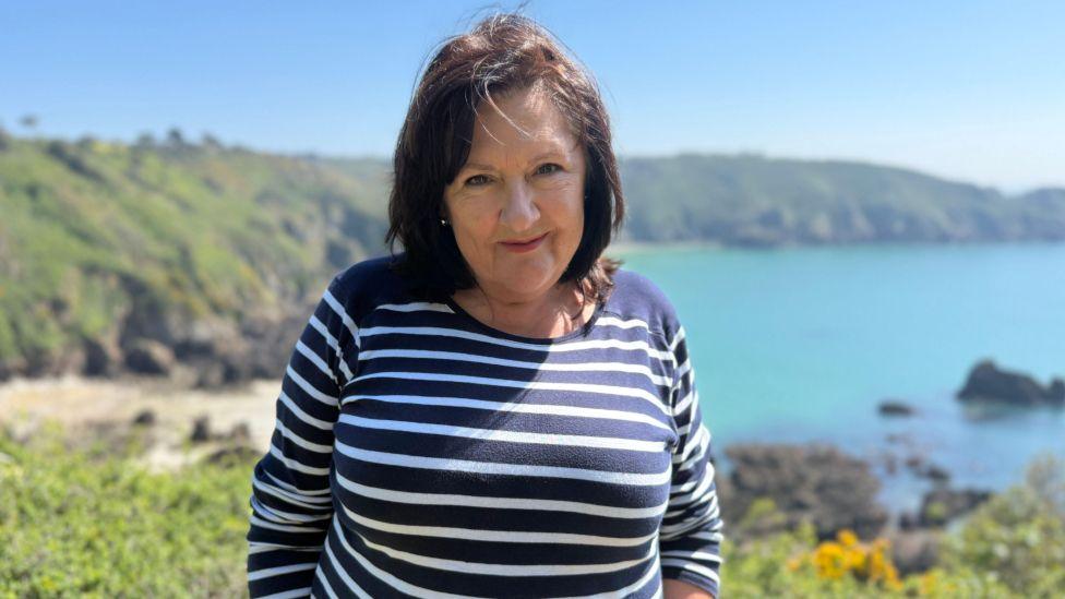 A lady with dark, chocolatey brown shoulder length hair looking at the camera smiling. She is wearing a Navy long sleeved top with thin white pin stripes. There are green cliffs and the blue sea in the background
