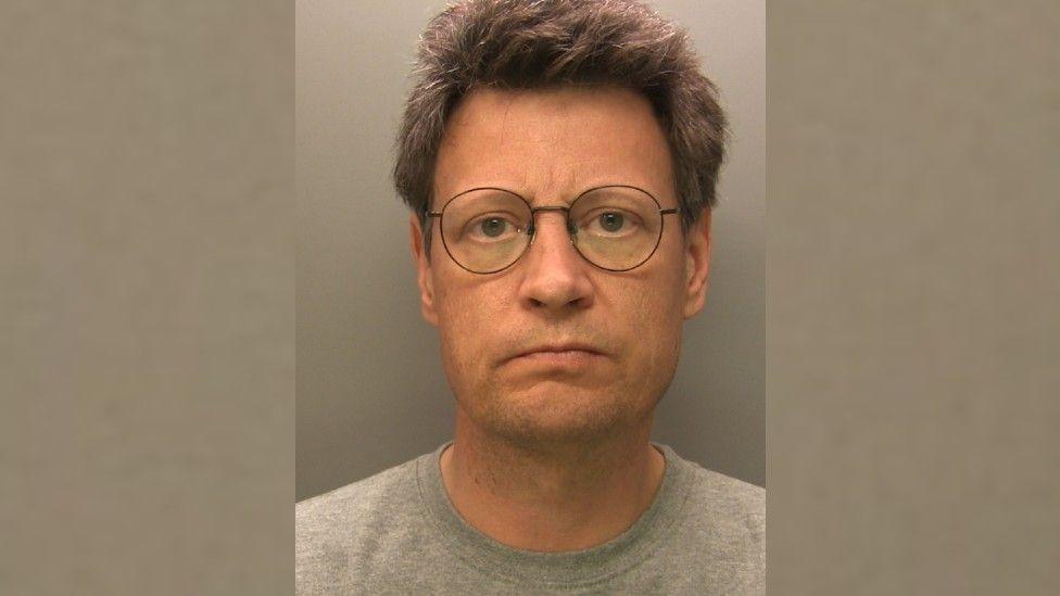 A mug shot of Daniel Holden. He has short grey hair and wears thin-rimmed black glasses and a grey t-shirt. 