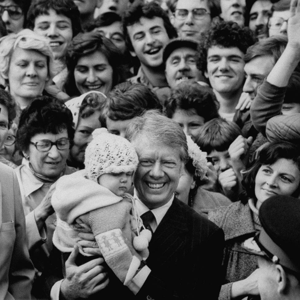 Former president Jimmy Carter hugging a baby, surrounded by a smiling crowd. The baby is wearing a knitted bonnet and jumper. Carter is wearing a suit and tie and is grinning widely.