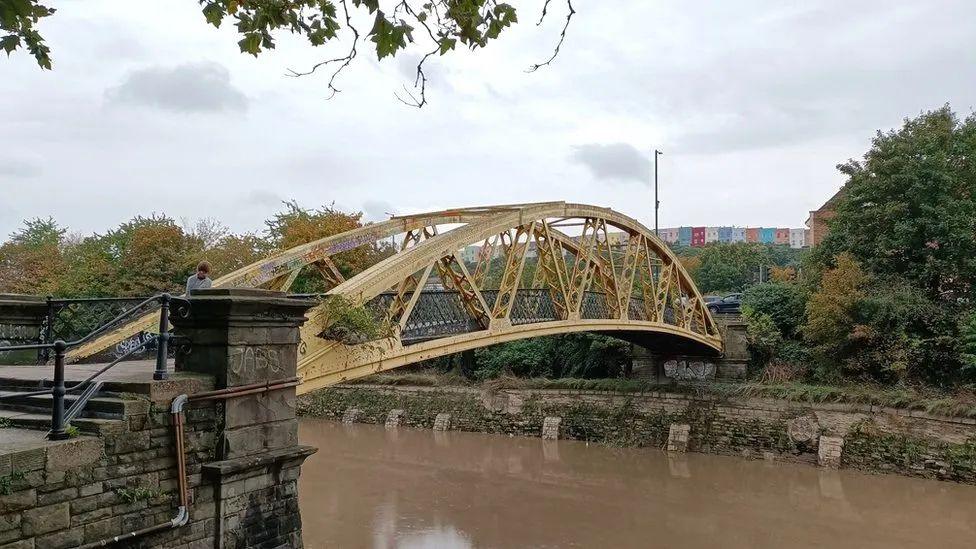 Langton Street Bridge in Bristol. It is yellow metal bridge spanning the river.