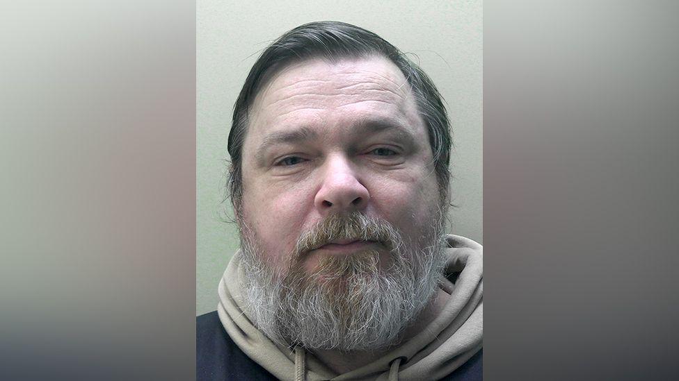 A headshot of a man with a grey beard wearing a beige hoodie underneath a black coat.