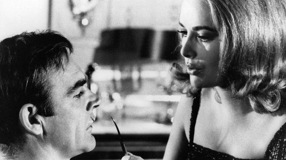 A black and white still from You Only Live Twice. On the left is Sean Connery, who has short hair and is in profile, with a cut lip. On the right is Karin Dor, who has shoulder-length thick blonde hair and is holding up a sharp implement.