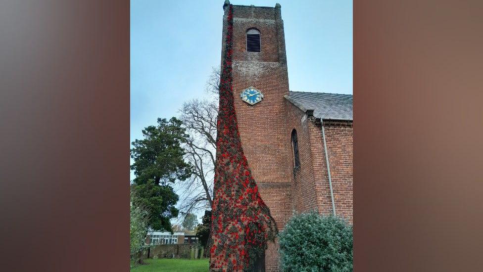 Thousands of knitted poppy flowers are attached to dark green netting, which is draped down the side of a red brick church tower.