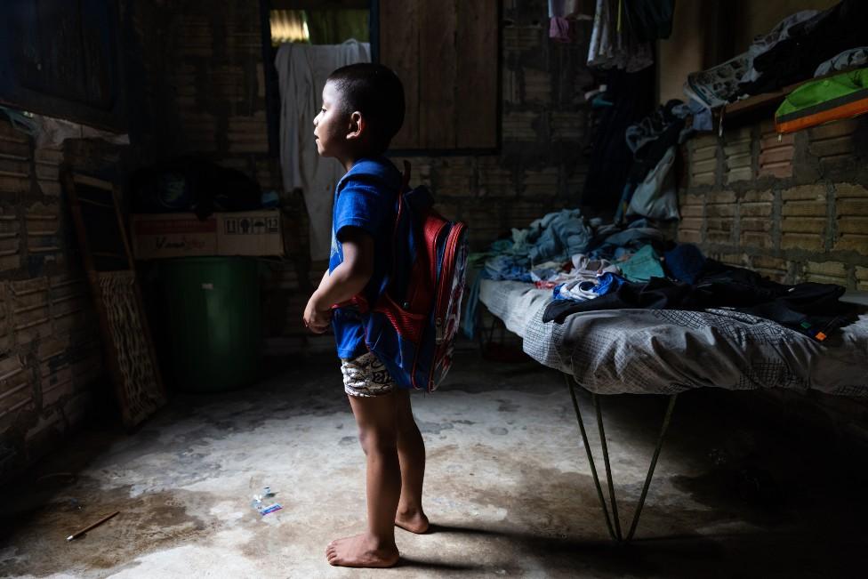 A child stands in a hut with his school backpack