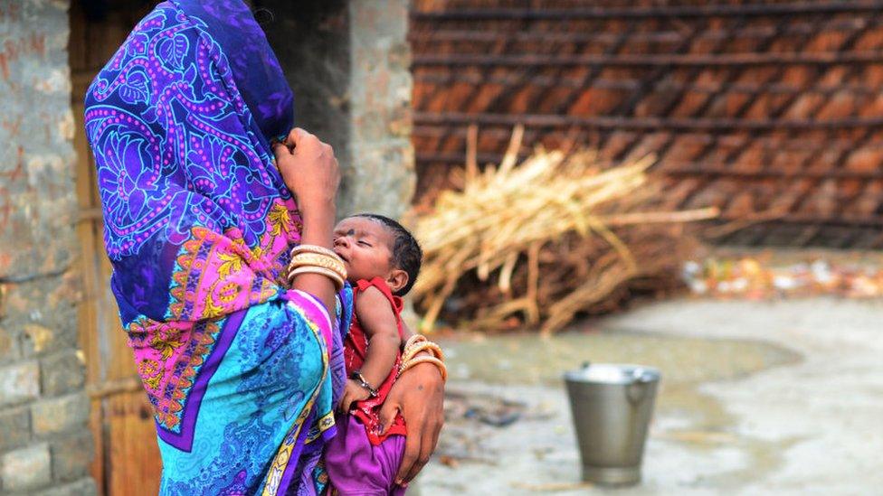 An Indian woman stands with her child, outside of her home in rural India.