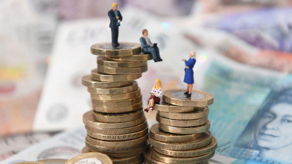 Models of men and women on a pile of coins and bank notes, illustrating the gender pay gap