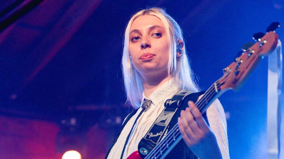 Georgia Davies from The Last Dinner Party. Georgia is a white woman in her 20s with long blonde hair, worn loose. She wears a white shirt buttoned to the top with a silver bolo tie. She also wears a fringed sleeveless leather jacket and has her bass guitar over her shoulder. She's pictured performing on stage in Austin, Texas, lit by red and blue lighting