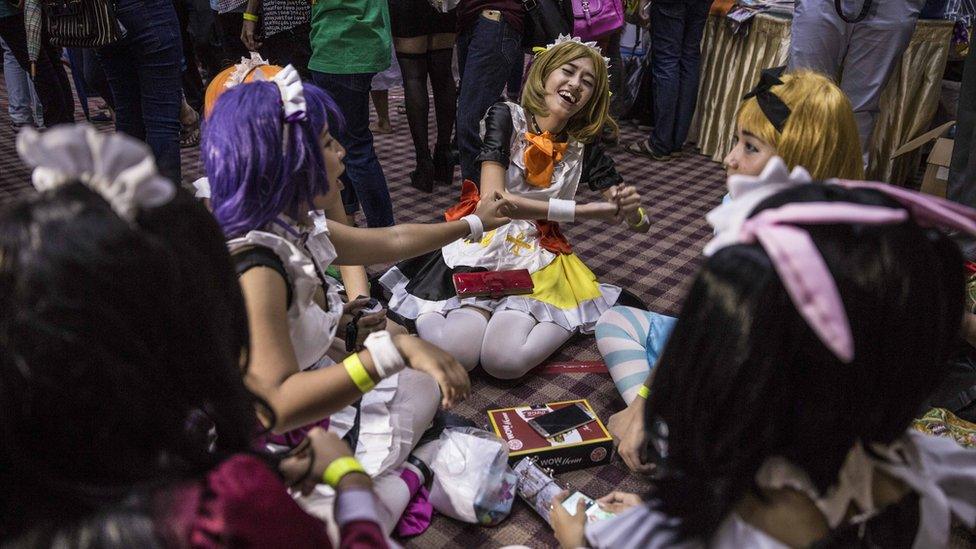 A group of five young women in cosplay, sitting in a circle and laughing, sitting on the floor of the main hall, with attendees standing around them