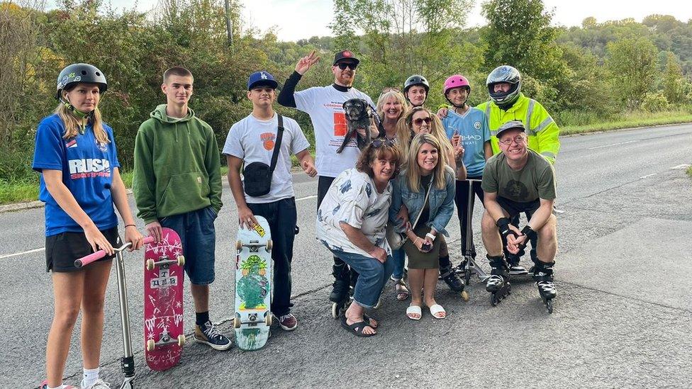 Si Coburn poses for a photograph with his dog, surrounded by several young people with skateboards in hand.