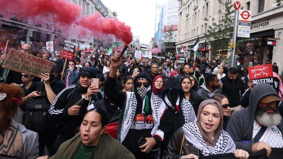 Protesters marching, one man with his face covered is holding a flare with red smoke pouring out of it