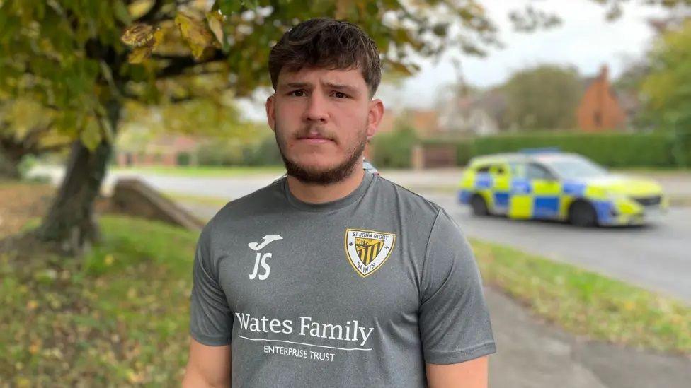 Joel Simpson looks at the camera, standing on the pavement with a grass verge either side and a tree to his right. In the background is a blurred police car, chequered neon yellow and blue. Joel wears a grey T-shirt with "Wates Family Enterprise Trust" on it and has brown styled hair and a beard